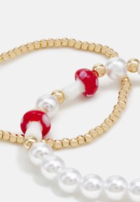 Pulseira de contas douradas com acentos de cogumelos brancos e vermelhos, misturada com pérolas brancas brilhantes e contas metálicas lisas. Elementos de design texturizados.