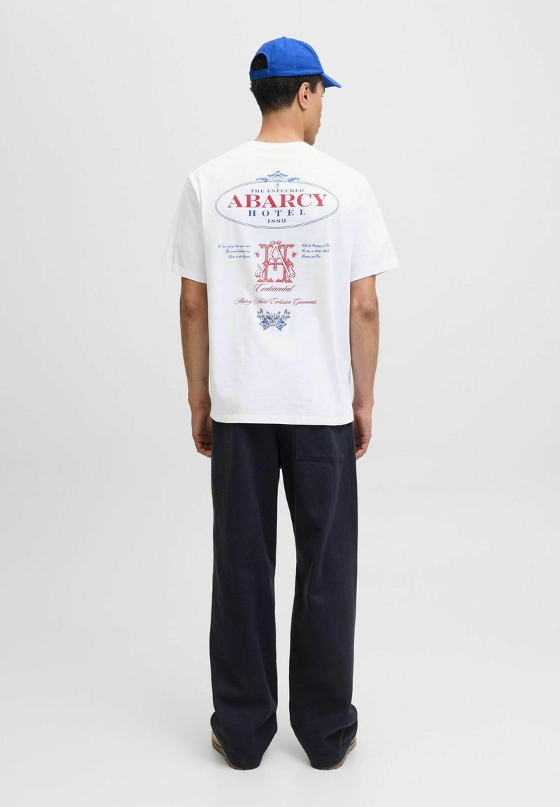Man wearing white graphic T-shirt with red and blue "Abarcy Hotel" text, navy pants, blue cap, standing facing away on plain background.
