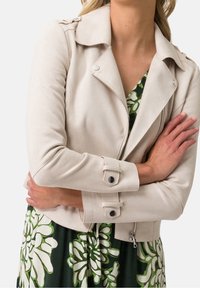 Beige motorcykeljakke i kunstlæder med lynlås, knapper ved manchetterne og en struktureret overflade, kombineret med en grøn blomsterkjole.