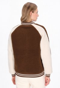 Brun og creme fleecejakke med raglanærmer, der har en ribbestrikket krave og manchetter med brune og hvide striber.