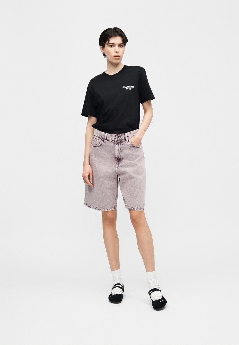 Junge Person trägt ein schwarzes Carhartt WIP T-Shirt, hellviolette Jeansshorts, weiße Socken und schwarze Slip-On-Schuhe, steht mit einer Hand in der Tasche.