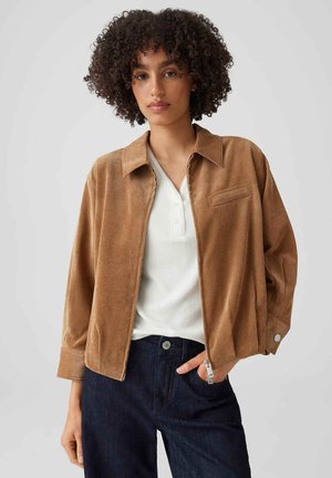Femme aux cheveux bouclés portant une veste marron, un haut blanc boutonné et un pantalon foncé, debout avec une main dans la poche devant un fond uni.