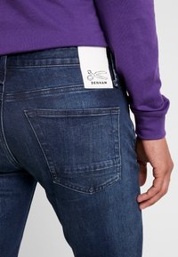Mörkblå denimjeans med en vit märkesetikett som har en saxlogotyp. Närbild av bakfickan och en hand som vilar nära midjan.