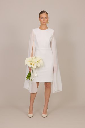 Γυναίκα με λευκό φόρεμα μέχρι το γόνατο με μακριά διάφανα μανίκια κάπα κρατά ένα μπουκέτο λευκές κάλλες, φορά λευκές γόβες και χρυσά σκουλαρίκια.