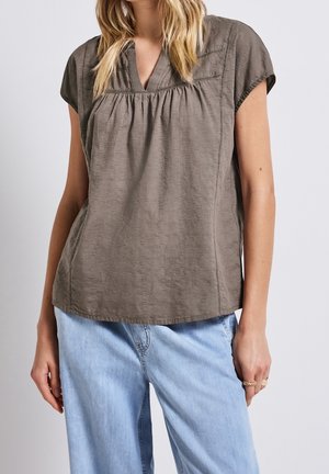 Vrouw draagt een losse bruine korte-mouw blouse met gestikte details en lichtblauwe spijkerbroek, staand tegen een effen achtergrond.