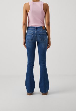 Personne portant un jean flare bleu et un haut côtelé rose sans manches, debout, de dos, devant un fond clair uni.