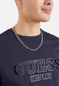 Camiseta marinera con "GUESS EST. 1981" impreso en letras negras brillantes; incluye un collar de cadena de metal plateado y un pequeño aro en la oreja.