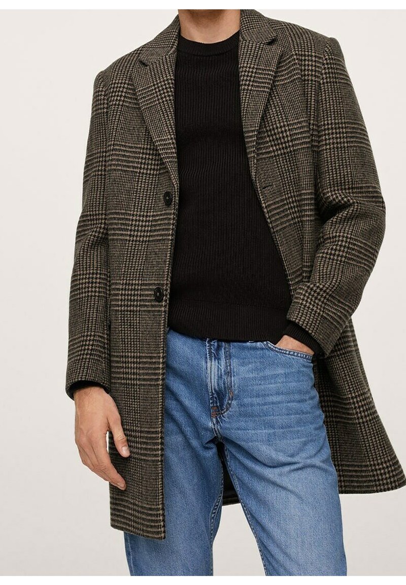 Homme portant un pardessus à carreaux marron et noir, un pull côtelé noir et un jean bleu clair, avec une main dans la poche et l'autre détendue.