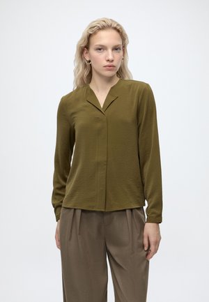 Frau mit blonden Haaren, die eine olivgrüne Bluse mit langen Ärmeln und braune plissierte weit geschnittene Hosen trägt, steht vor einem weißen Hintergrund.