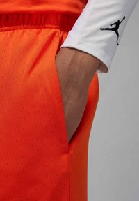 Lys oransje treningsshorts med et teksturert mønster, med en sidelomme og en hvit langermet topp med en svart logo.