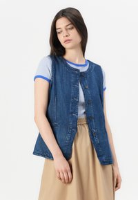 Chaleco de denim con escote redondo, cierre frontal de botones y dos bolsillos laterales. Combinado con una camiseta de rayas y una falda beige.