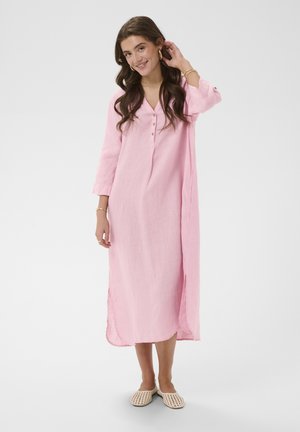 Femme aux longs cheveux bruns portant une robe midi rose ample avec des boutons et des chaussures plates en toile tressée crème, debout devant un arrière-plan uni.