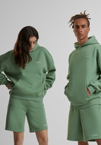 Deux personnes portent des sweat-shirts à capuche verts assortis, oversize, avec des shorts, comprenant une poche avant et un logo brodé. Le matériau semble doux et texturé.