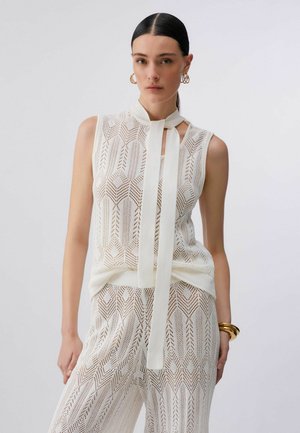 Donna con capelli scuri indossa un top bianco senza maniche lavorato a maglia con cravattino e pantaloni a gamba larga abbinati, accessoriata con orecchini a cerchio dorati e braccialetti.