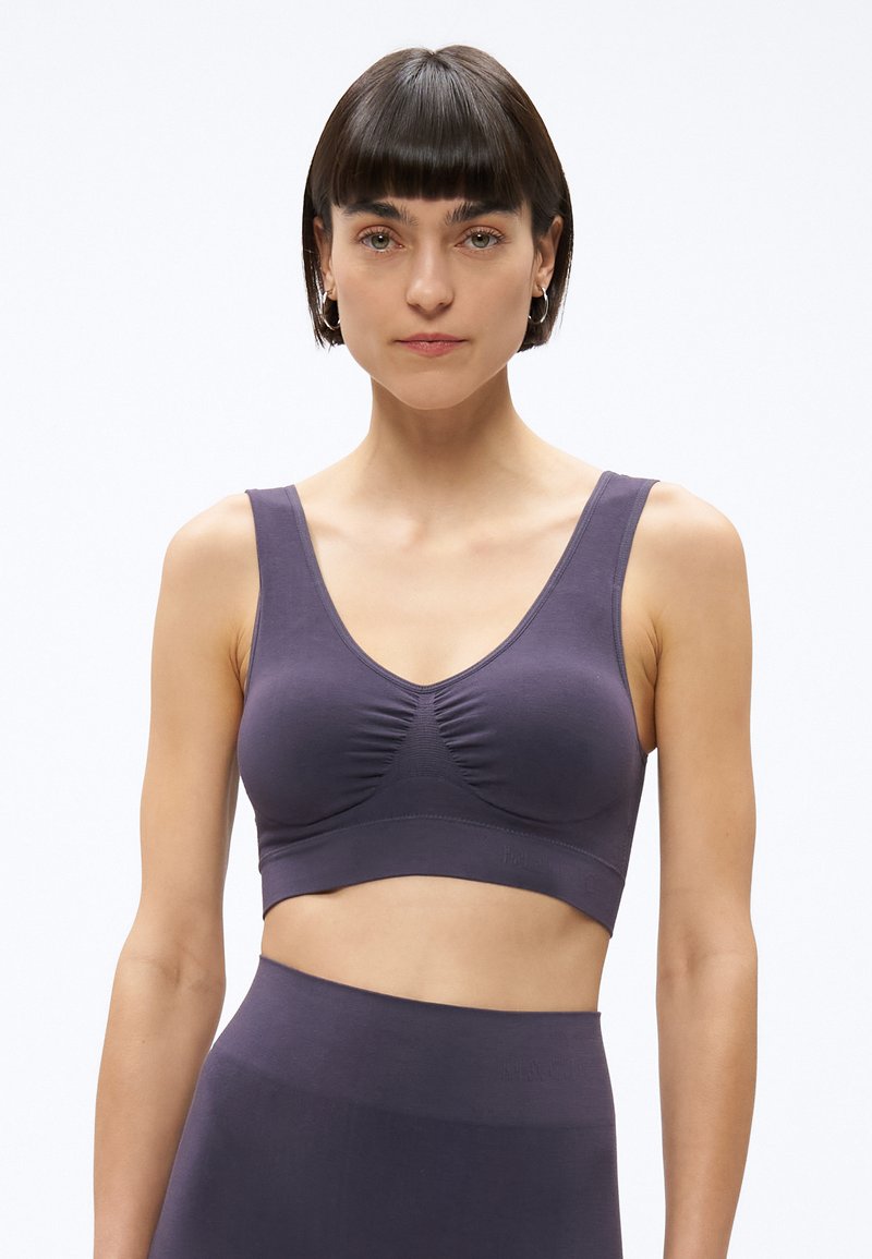 Žena s krátkymi tmavými vlasmi, oblečená v fialovom crop tope bez rukávov a zodpovedajúcich vysokými pásovými nohavicami, stojaca na jednoduchom svetlom pozadí.