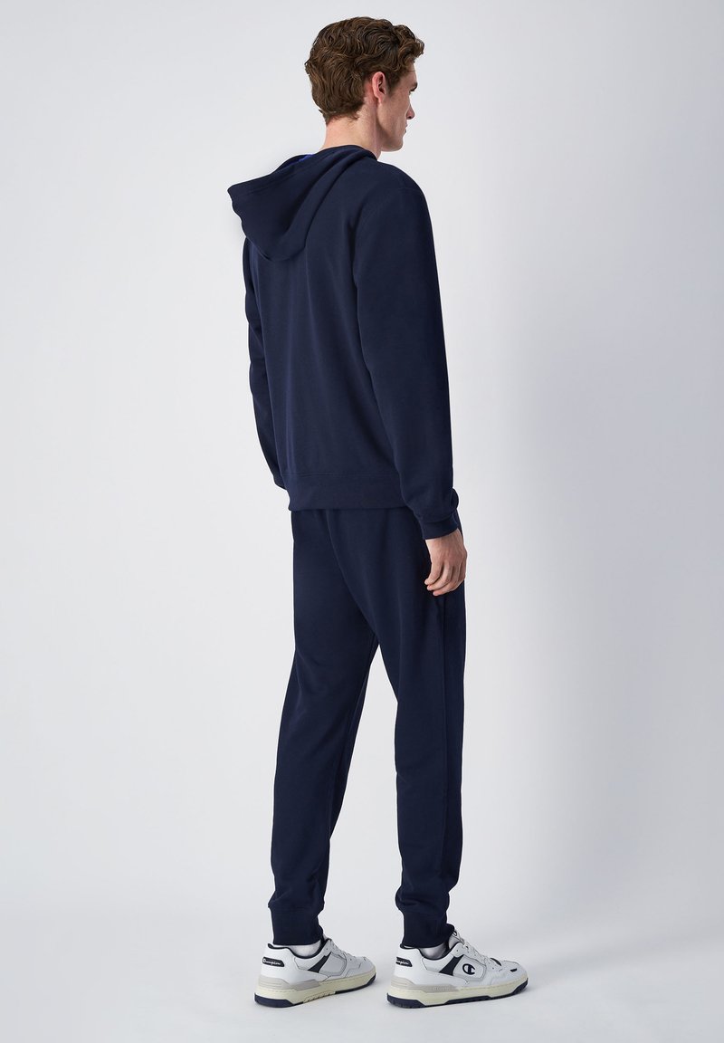 Marineblauwe hoodie en bijpassende joggers gemaakt van zachte stof, met een trekkoordcapuchon, ribgebreide manchetten en sneakers met witte en zwarte accenten.
