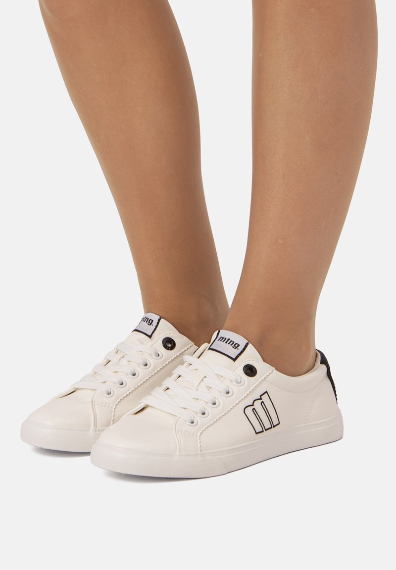 Zapatillas blancas con cordones hechas de un material similar al cuero, con un detalle negro en el talón y un logo minimalista en el lateral. Suela de goma plana.