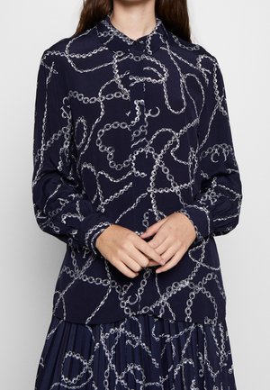 Femme portant un chemisier bleu marine et une jupe plissée avec un motif de maillons de chaîne blancs, boutonnée au col, mains doucement jointes.