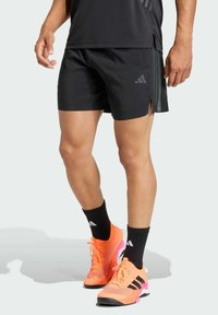 Pantalones cortos deportivos negros con aberturas laterales, combinados con zapatillas deportivas naranja brillante con detalles en negro y rosa, y calcetines tobilleros negros con logotipos.