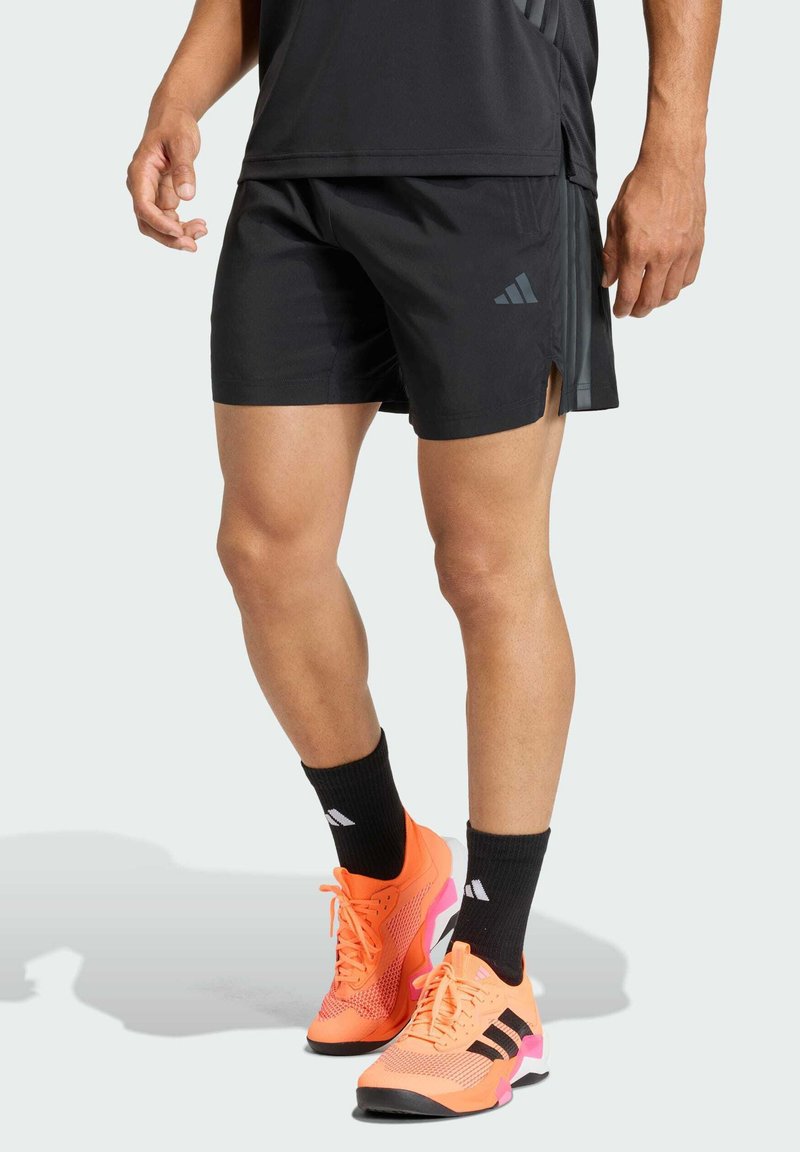 Pantalones cortos deportivos negros con aberturas laterales, combinados con zapatillas deportivas naranja brillante con detalles en negro y rosa, y calcetines tobilleros negros con logotipos.