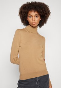 Beige garbónyakú pulóver sima kötött anyagból, hosszú ujjakkal és slim fit szabással. Egyenes alj, oldalán finom varrással.