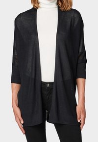 Svart stickad kofta med trekvartslånga ärmar, öppen fram och lätt textur, lager på lager över en vit polotröja.