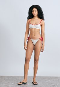Σετ μπικίνι σε λευκό με σχέδιο πλέγματος, διακοσμημένο με κόκκινο περίγραμμα. Το πάνω μέρος έχει δομημένο σχέδιο, ενώ το κάτω μέρος διαθέτει ρυθμιζόμενους δεσμούς στα πλευρά.