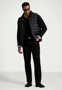 Muž nosi čierne slnečné okuliare, čiernu puffy vestu cez čierny sveter, čierne nohavice a čierne čižmy, stojí s jednou rukou v vrecku.