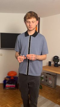 Siva polo majica kratkih rukava s crnom ovratnikom i plaketom za gumbe. Model drži narukvicu, pokazujući tetovaže na rukama. Tamne hlače.