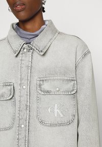 Svetle šedá džínová bunda s límcem, dvěma předními kapsami, kovovými patentkami a vyšitým logem "CK" na levé kapse.