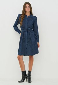 Sötétkék farmer ruha hosszú ujjakkal, gombos rögzítéssel, összehúzott elülső résszel, övvel szabott derékkal és térdig érő szegéllyel. Mellette szegecselt fekete bokacsizma párosítva.