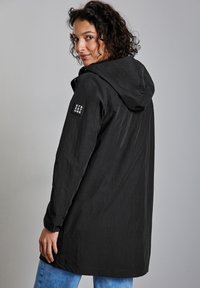 Czarna kurtka z kapturem o teksturowanej tkaninie, z długimi rękawami i logo na lewym ramieniu. Charakteryzuje się prostym krojem i sięga do połowy uda. Widać niebieskie dżinsy.