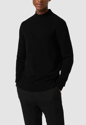 Black knitted sweater with a high neck, ribbed cuff, and hem. Soft texture, fitted silhouette. Paired with dark trousers.