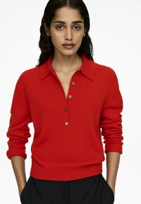 Roter gestrickter Polo-Pullover mit Kragen, drei braunen Knöpfen und gerippten Bündchen. Der Stoff wirkt weich mit einer glatten Textur.