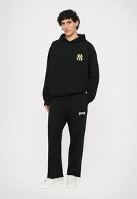 Čierna mikina s kapucňou a logom na ľavej hrudi, v kombinácii s čiernymi teplákmi so bielym textom. Hladká tkanina, voľný strih a detaily kapuce.