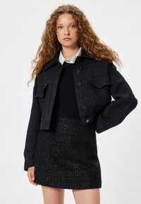 Musta lyhennetty takki, jossa on rakenteellinen kangas, kaksi rintataskua ja mustat napit, yhdistettynä vastaavaan mustaan tweed-minihameeseen.