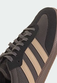 Zapatilla negra y marrón con una parte superior texturizada, cordones grises y tres rayas beige a lo largo del lateral. Suela de goma con borde dentado.