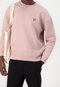 Man in een lichtroze sweatshirt met een geel vogellogo, zwarte broek, met een beige canvas boodschappentas over zijn schouder.