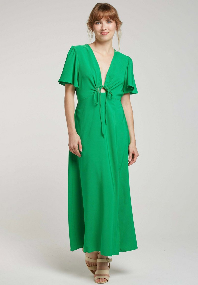 Robe maxi verte avec un décolleté en V profond, un détail de taille nouée, des manches flottantes et un tissu fluide. Portée avec des sandales à plateforme de couleur claire.