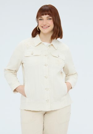 Smilende kvinde med rødt hår iført cremefarvet, knappet jakke og matchende bukser, hænderne i lommerne, foran en ensfarvet lys baggrund.