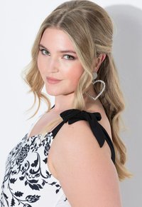 Abito floreale in bianco e nero con dettagli a fiocco sulle spalline, che evidenzia un design aderente. La modella presenta capelli biondi ondulati e grandi orecchini a forma di cuore.