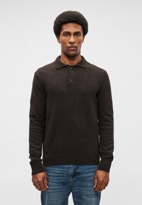 Καφέ πλεκτό πουλόβερ με πόλο γιακά, μπροστινά κουμπιά και ριγέ μανσέτες, σε συνδυασμό με μπλέ τζιν παντελόνι. Υφασμάτινη υφή σε σκούρο χρώμα με υποψία μοτίβου.