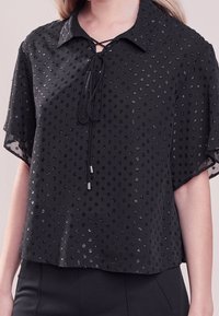 Blouse noire à manches courtes avec un col, un décolleté lacé et des points texturés avec une finition brillante. Tissu doux et léger avec une coupe ample.