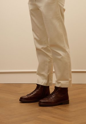 Bottes en cuir marron avec un laçage à l'avant, semelle surélevée et bout rond. Associées à un pantalon blanc, ajusté et léger sur un sol en bois.