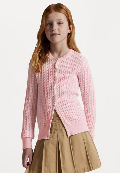 Cárdigan de punto color rosa con puños y dobladillo acanalados, que presenta botones amarillos y un diseño dividido. Llevado sobre una falda plisada color arena.
