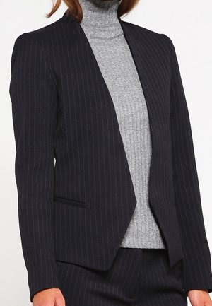 Kvinde iført en sort blazer med fine striber over en grå ribstrikket rullekravesweater, kombineret med matchende buks med fine striber.