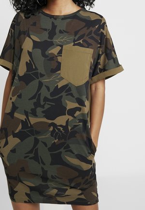 Kvinde iført en kjole med camouflage mønster med opkrogede korte ærmer og en ensfarvet olivengrøn brystlomme.