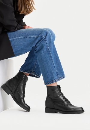 Czarne skórzane botki za kostkę z gładką powierzchnią, sznurowane z przodu i z bocznym zamkiem, zestawione z podwiniętymi niebieskimi dżinsami. Kwadratowy obcas.