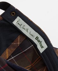 O rótulo de algodão apresenta "Paul Smith ama Barbour" em texto verde, colocado contra um interior estampado de xadrez azul escuro e castanho com detalhes laranja.