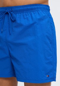 Μπλε ανδρικό μαγιό με ελαστική μέση, κορδόνι και μικρό λογότυπο κοντά στο τελείωμα, φορεμένο απευθείας πάνω στο δέρμα.